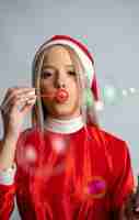 Free photo young fair-haired female posing in a miss santa claus costume and blowing bubbles