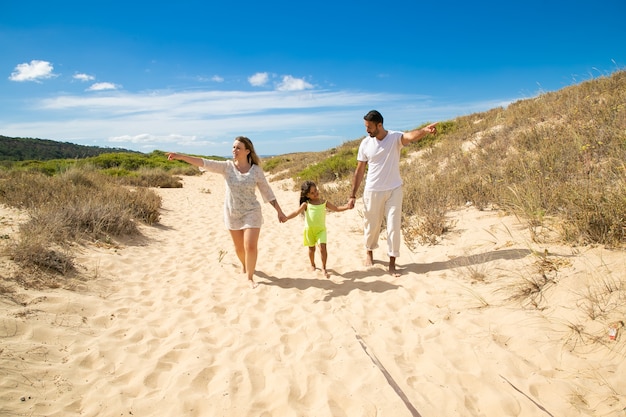 Young family couple and little kid in summer clothes walking white along sand path, pointing hands away, girl holding parents hands