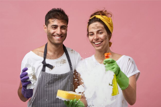 Coppia giovane famiglia lavare la superficie del vetro con un detergente che pulisce la schiuma gioendo dei buoni risultati della pulizia essendo soddisfatto dello spray di lavaggio. sporchi lavoratori maschi e femmine che puliscono le finestre a casa