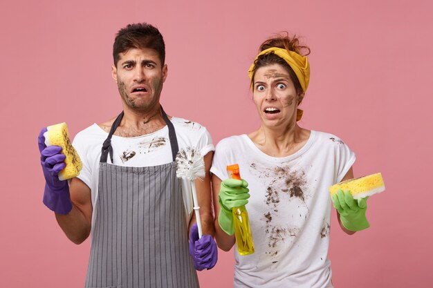 Young family couple with dirty faces having disgusting look holding window cleaner and sponges washing windows in living room. Male and female completing their work about house having bad mood