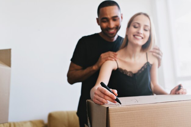 Young family moving to new house, buy apartment, flat. Cheerful couple packing boxes with books, writing labels. They in white room with window, wearing black top and T-shirt.