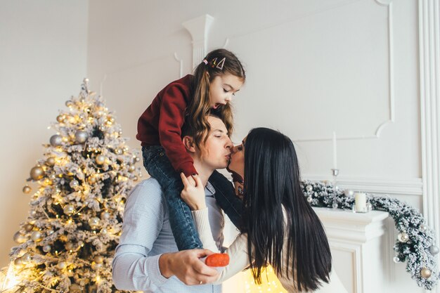 Young family poses before a shiny Christmas tree in a cosy luxury room