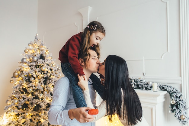 若い家族は居心地の良い豪華な部屋の光沢のあるクリスマスツリーの前にポーズを取る
