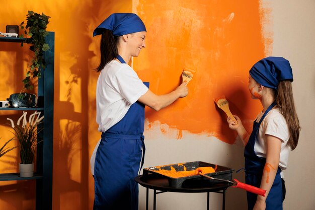 Young family using brush to paint walls with orange color and renovating tools, having fun with messy redecoration work. Adult with little messy girl decorating apartment interior with paintwork.