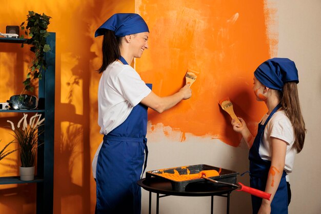 젊은 가족은 브러시를 사용하여 주황색으로 벽을 칠하고 도구를 수리하고 지저분한 재장식 작업을 즐깁니다. 페인트칠로 아파트 내부를 장식하는 지저분한 소녀를 둔 성인.