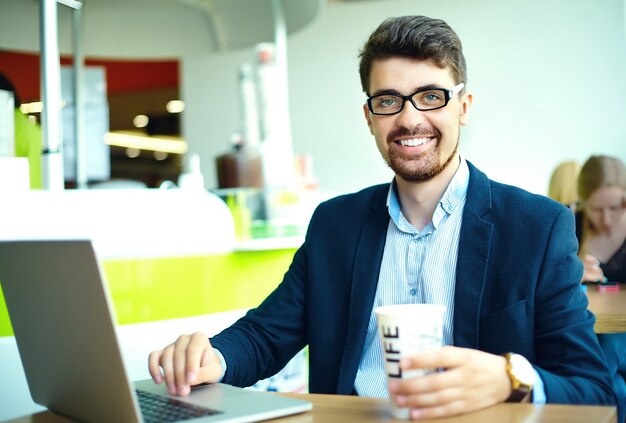 Молодой модный улыбающийся хипстерский человек, пьющий кофе в городском кафе во время обеда с ноутбуком в костюме