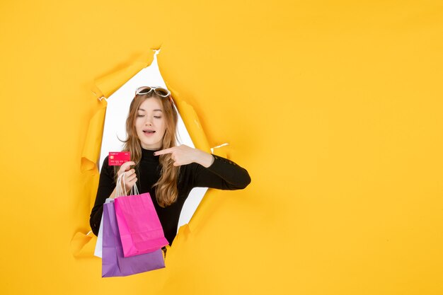 Young fashion woman holding shopping bags and credit card through torn paper hole in the wall