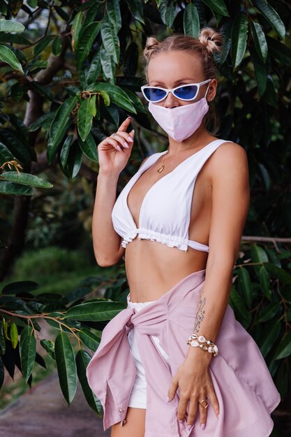 Young fashion woman in white crop top, sunglasses and white medical face mask in the nature