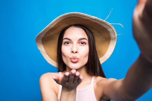 Young fashionable attractive girl in sunglasses and straw hat sends air kiss to camera. Nice playful and happy woman with red lips on blue wall. with copy space.