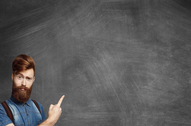 Young fashionable man with fuzzy beard wearing denim shirt and suspenders looking surprised or confused while pointing his index finger at blank chalkboard with copy space for your advertising content