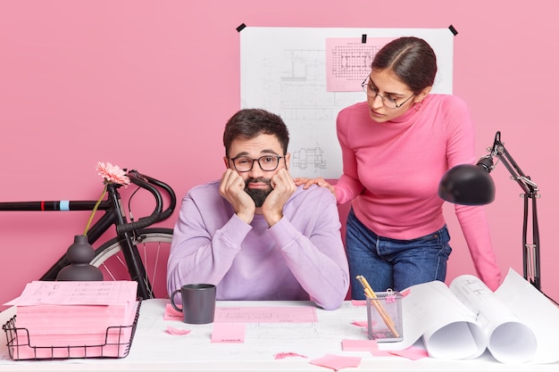 Young female architect and male engineer collaborate together for common project. Sad bored bearded man poses in creative office with colleague tired of working on blueprints. Teamwork concept