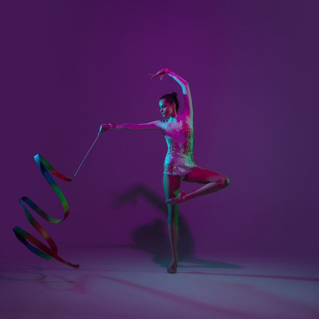 Young female athlete, rhythmic gymnastics artist dancing, training isolated on purple studio background with neon light. Beautiful girl practicing with equipment. Grace in performance.