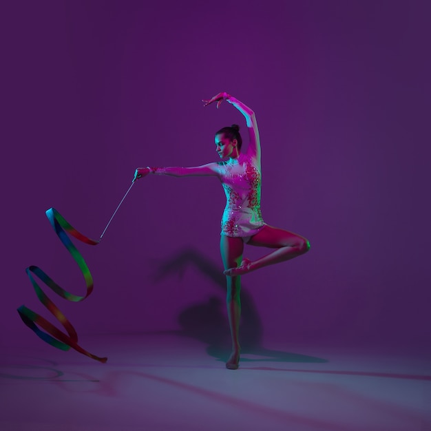 Молодая спортсменка, художник по художественной гимнастике танцует, тренируется на фиолетовом фоне студии с неоновым светом. Красивая девушка тренируется с оборудованием. Изящество в исполнении.