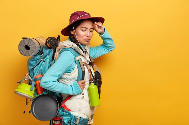 Young female backpacker has tired face expression, keeps hand on forehead, feels fatigue after long trip on foot