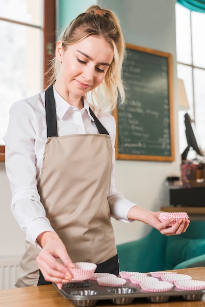 Young female baker placing an empty paper forms for muffins