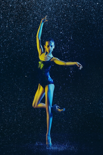 Young female ballet dancer performing under water drops and spray. Caucasian model dancing in neon lights. Attractive woman. Ballet and contemporary choreography concept. 