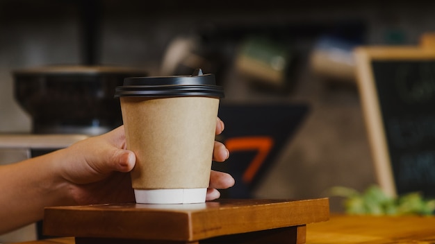 Young female barista serving take away hot coffee paper cup to consumer standing behind bar counter at cafe restaurant.