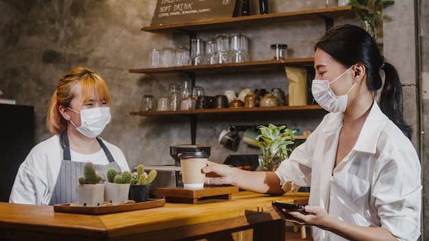 젊은 여성 바리 스타는 카페에서 소비자에게 뜨거운 커피 종이 컵을 가져가는 얼굴 마스크를 착용합니다.