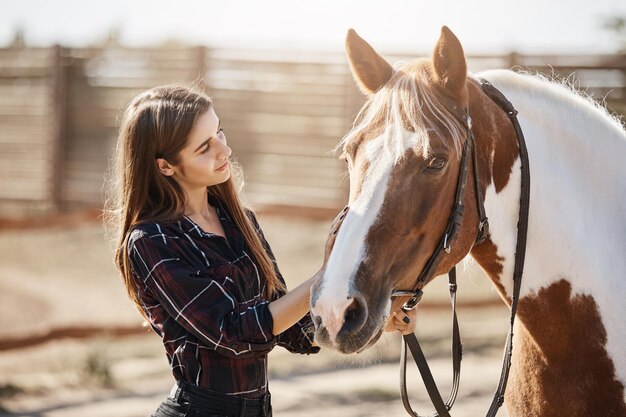 Young female barn manager taking care and talking to a young stallion preparing for a ride