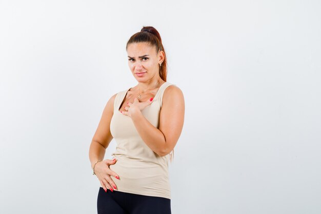 Young female in beige tank top holding hand on chest and looking hesitant