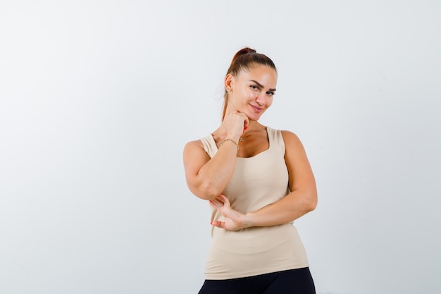 Young female in beige tank top holding hand under chin and looking cute , front view.