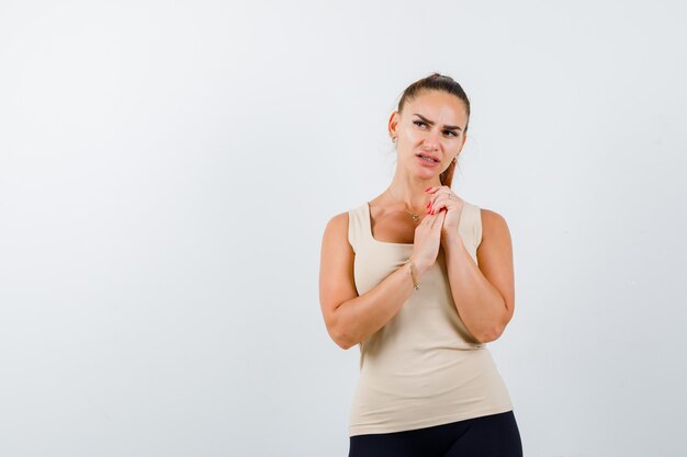 Young female in beige tank top holding hands together over chest and looking pensive , front view.