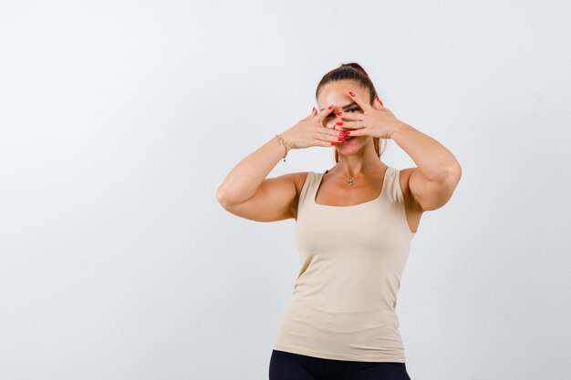 Young female in beige tank top looking through fingers and looking cute , front view.