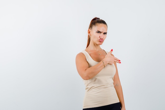 Free photo young female in beige tank top showing gun gesture and looking confident , front view.