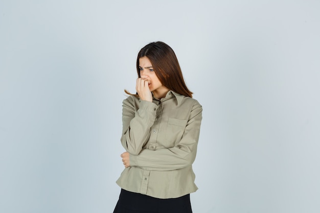 Young female biting nails emotionally in shirt, skirt and looking anxious. front view.