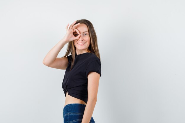 Young female in black blouse, pants showing ok sign on eye and looking joyful