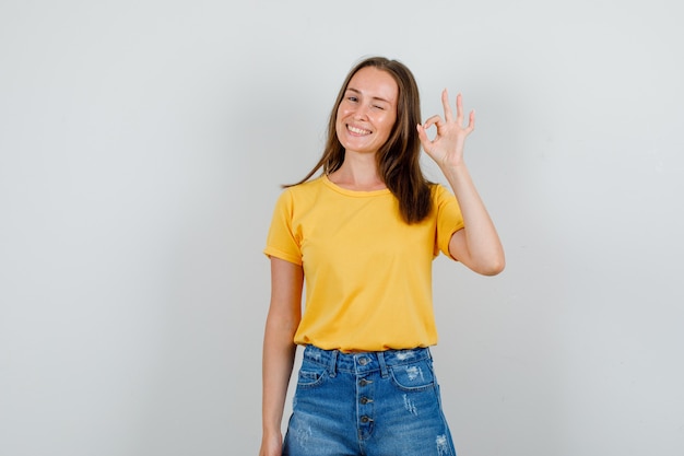 Young female blinking eye with ok sign in t-shirt, shorts and looking cheerful