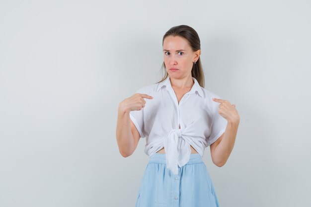 Young female in blouse, skirt pointing at herself and looking confused