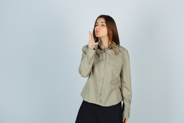 Young female blowing on finger pistol with pouted lips in shirt, skirt and looking confident. front view.