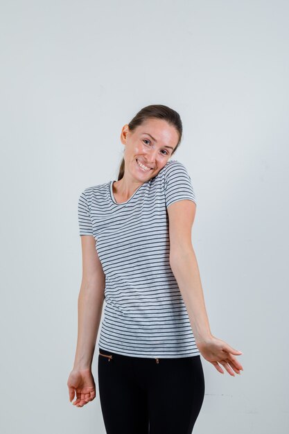 Young female bowing head on shoulder in t-shirt, pants and looking optimistic , front view.
