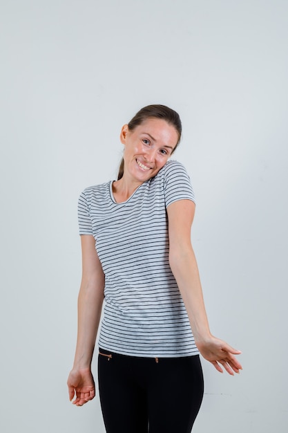 Young female bowing head on shoulder in t-shirt, pants and looking optimistic , front view.