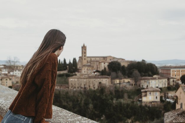 Молодая женщина в коричневом свитере стоит на мосту и наслаждается прекрасным видом на городской пейзаж