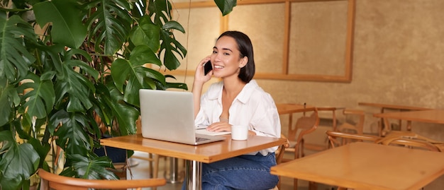 젊은 여성 카페 매니저 소유자가 노트북과 함께 앉아 전화 통화에 응답하고 우호적으로 미소 짓고 있습니다.