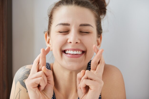 Young female Caucasian student keeping her fingers crossed and eyes closed to make a desired wish. Charming white smile, positive heartwarming look of girl sincerely believing in her victory.