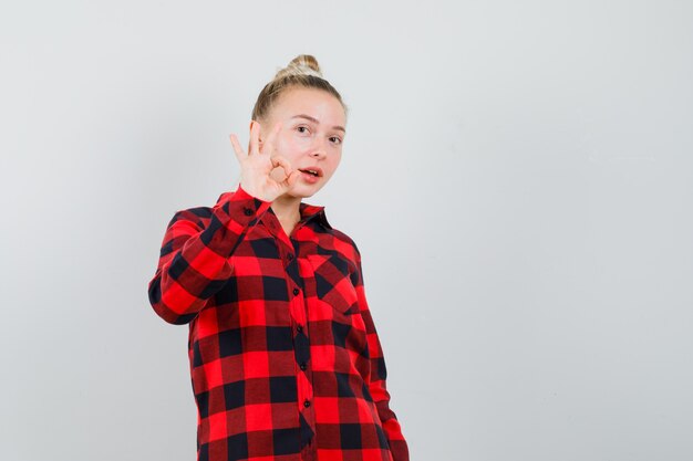 Young female in checked shirt doing ok gesture and looking confident , front view.