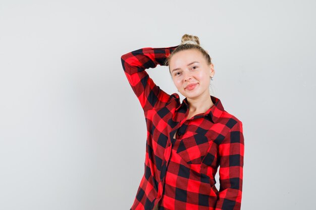 Young female in checked shirt holding hand behind head and looking cheerful , front view.