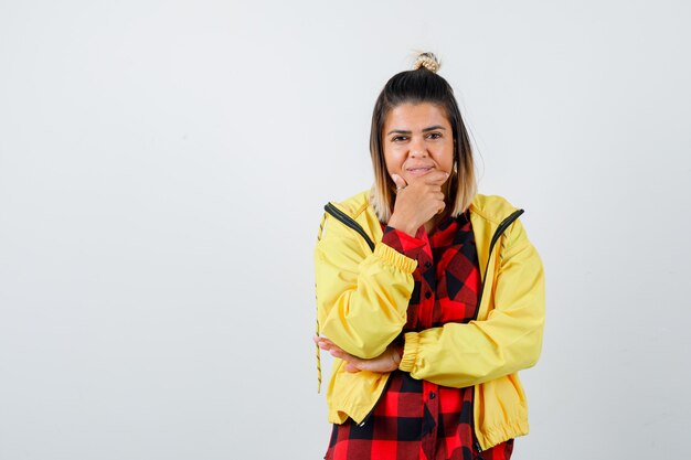 Young female in checkered shirt, jacket holding fingers on cheeks and looking merry , front view.