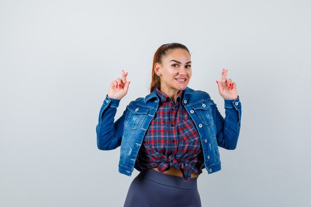 Young female in checkered shirt, jacket, pants showing crossed fingers and looking joyous , front view.