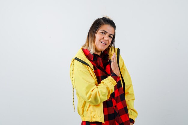 Young female in checkered shirt, jacket showing something with spreading palm and looking cheerful , front view.