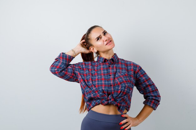 Young female in checkered shirt, pants with hand behind head while keeping hand on hip and looking pensive , front view.