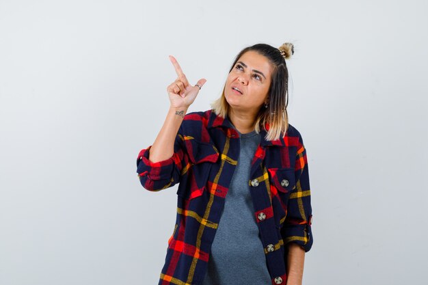 Young female in checkered shirt pointing at upper left corner and looking confident