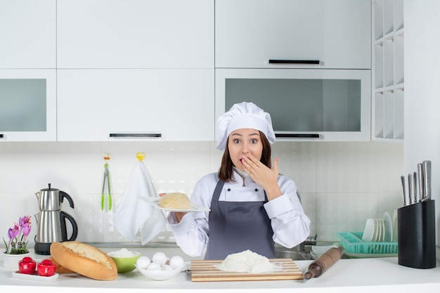 Молодая женщина-шеф-повар в униформе стоит за столом с едой на разделочной доске и держит готовую еду с удивленным выражением лица на белой кухне