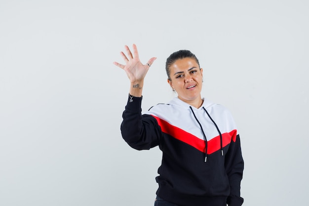 Young female in colorful sweatshirt waving hand for greeting and looking pleased , front view.