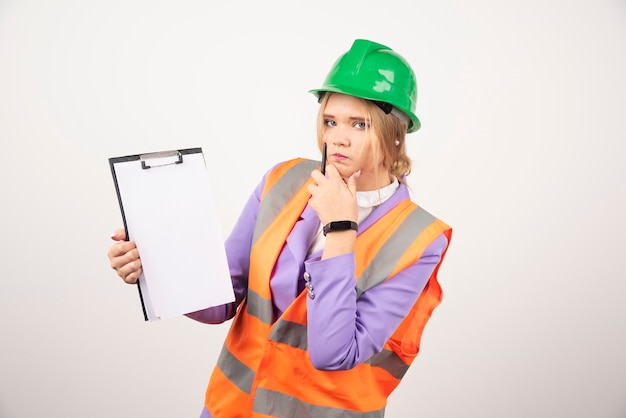 Young female contractor with green helmet and clipboard on white wall. 