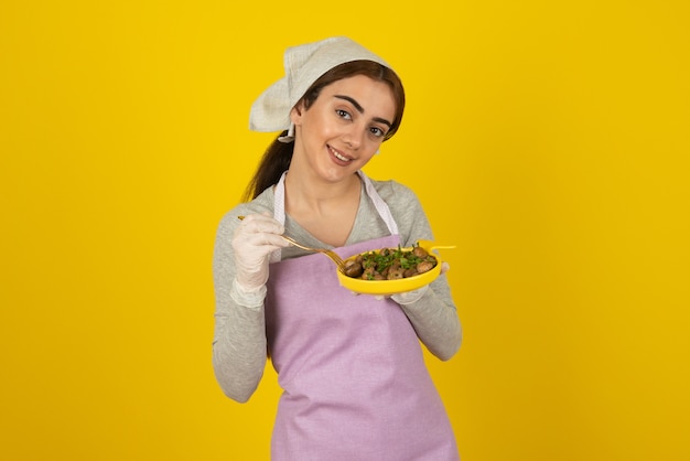 黄色い壁に揚げたキノコを食べるエプロンで若い女性料理人。