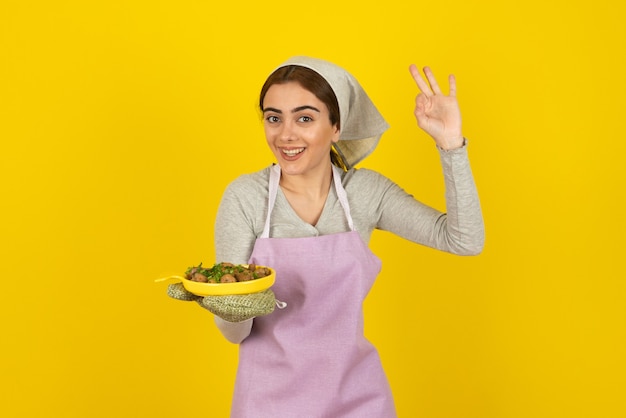 Young female cook in apron holding plate of fried mushrooms and showing ok sign.